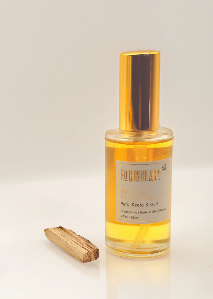 A clear glass bottle of Fumulary Palo Santo & Oud diffuser oil filled with golden yellow liquid, topped with a shiny gold cap, shown on a white background with a Palo Santo wood stick beside it.