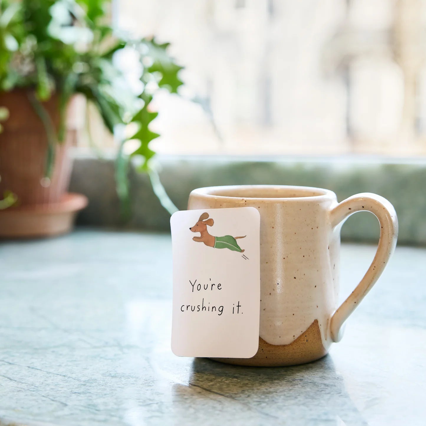 Ceramic mug with a motivational card on a table with plants in the background